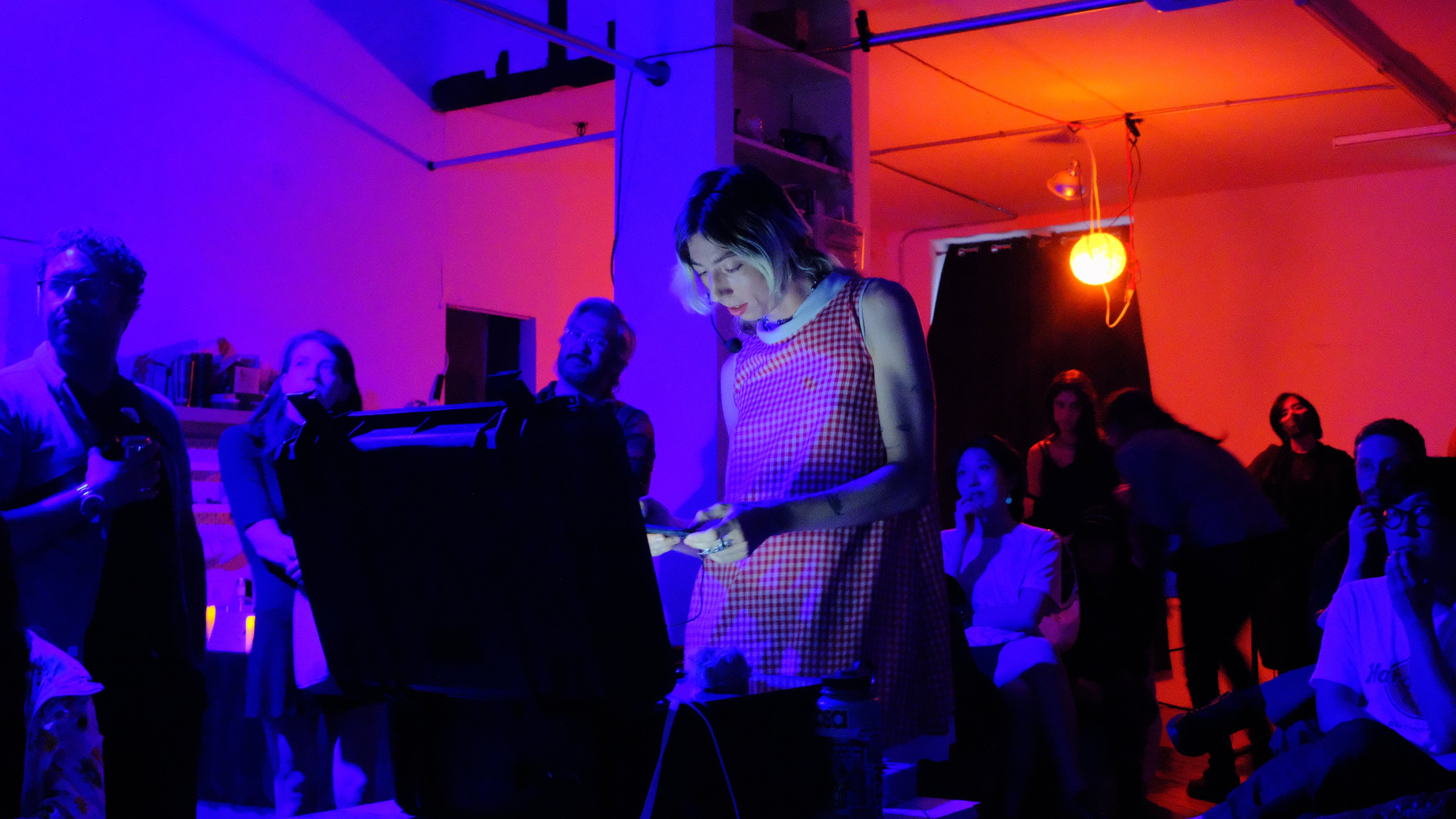A trans woman (me) wearing a red gingham a frame dress stands smiling behind an open Pelican case in a dark room surrounded by people and lit in dramatic blue.