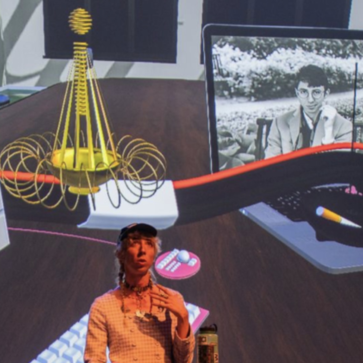 A trans woman (me) with long thin braids in a lavender tweed skirt suit standing behind a lectern with a projection of a virtual world behind her. On the left of the projection is a gold chandelier looking shape (my avatar) and on the right is a laptop with a portrait of a dour young man (my dead husband) on the screen.