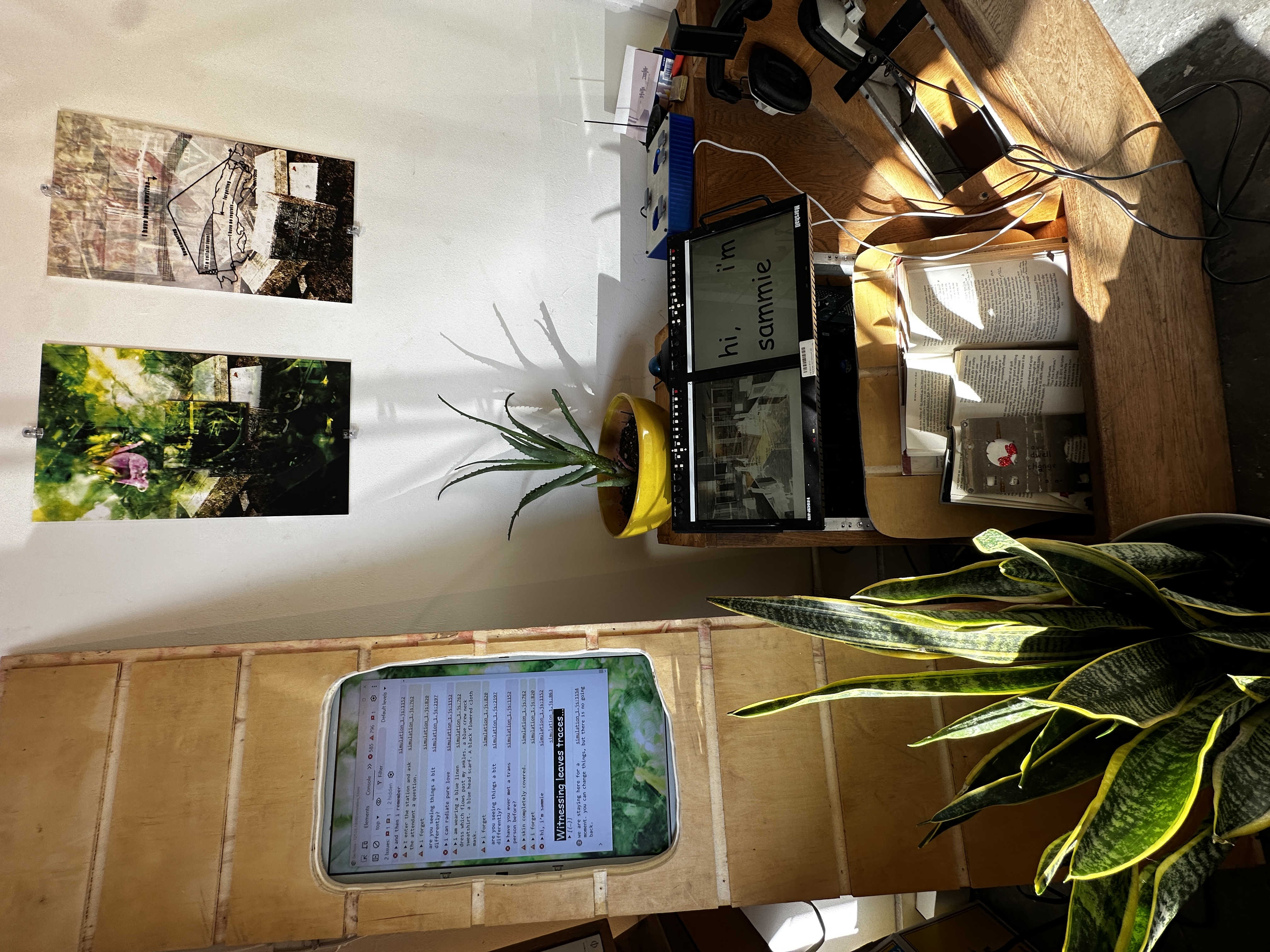 a strange wooden computer surrounded by plants. A controller with four buttons sits on top, and a pair of closed back studio headphones hang on a hook. the vertical monitor leaning against the wall is made from a 6 foot board with a wavy rectangle cut out and an old LCD mounted behind.  it displays a browser console full of warnings and errors. the main computer case has a dual rackmounted monitor unit at about waist level, with two open books resting vertically in the nook beneath. The left monitor displays a chaotic multilayered image. the right displays the text ‘hi i’m sammie’. 