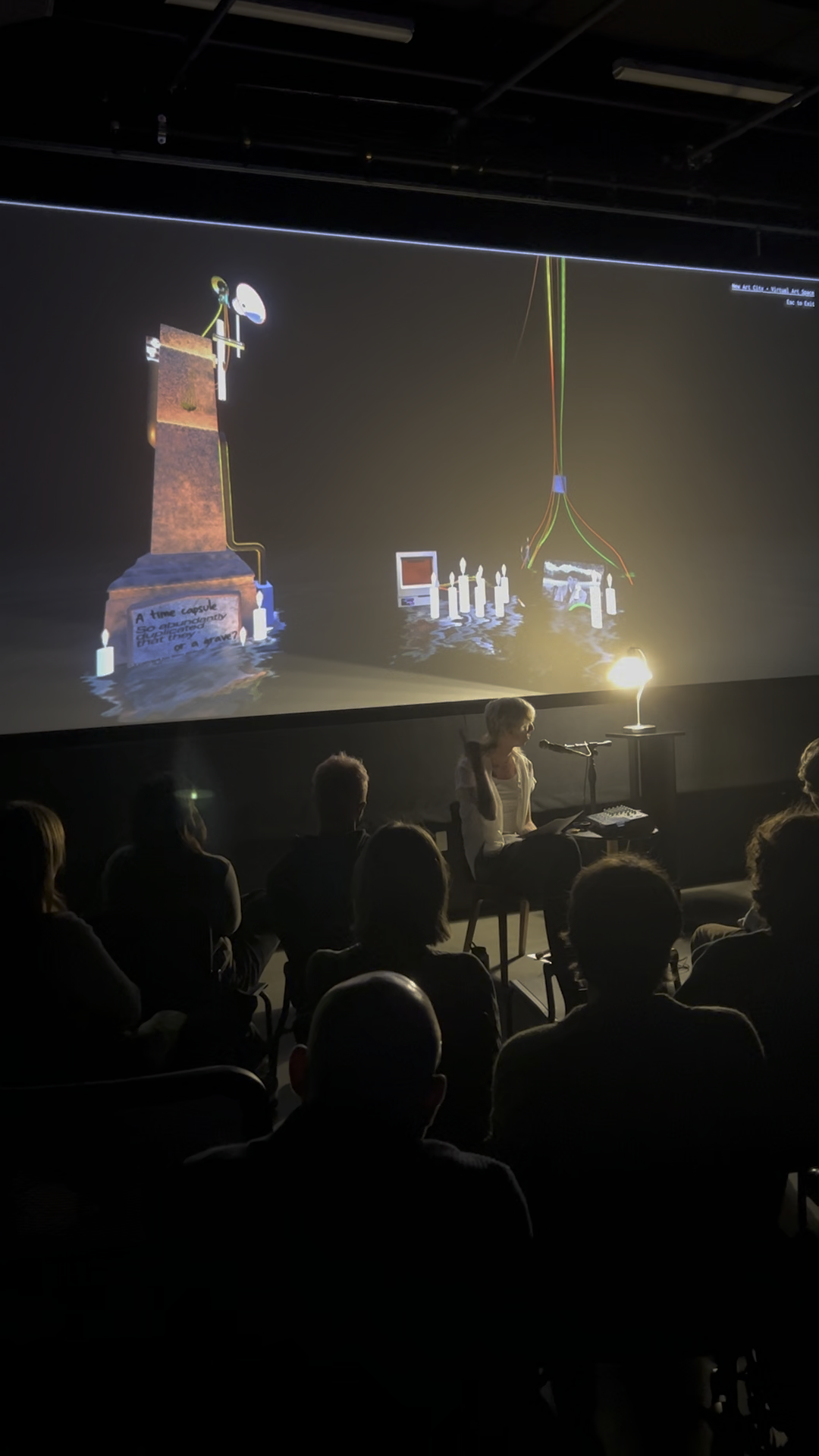 A trans woman (me) wearing a tank top and open short sleeved button-up seated in a dark black box theater with a projection behind her of a memorial obelisk sitting in dark water with a computer altar sitting on a rock beside it.